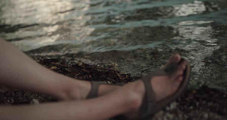 Woman wearing sandals sits next to the lake as the small waves roll in
