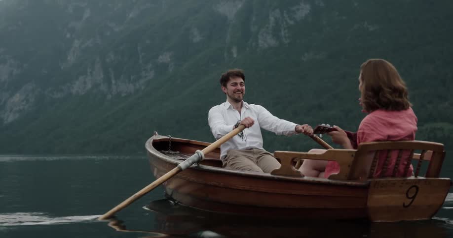 A man and woman are in a small boat on a lake near a mountain and he is rowing as she plays a ukulele