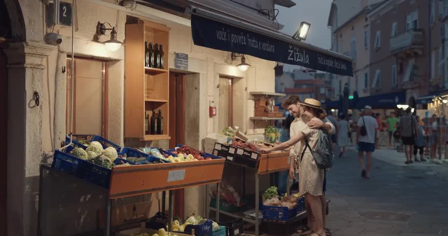 Husband and wife approach a fruit and vegetable market and the woman feeds the man some produce