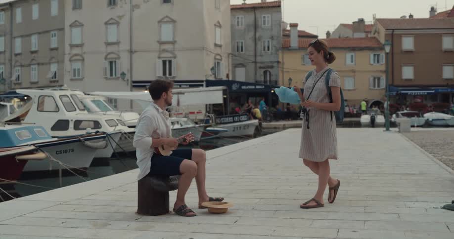 Woman eats cotton candy while listening to a man play the ukulele next to the boat harbor