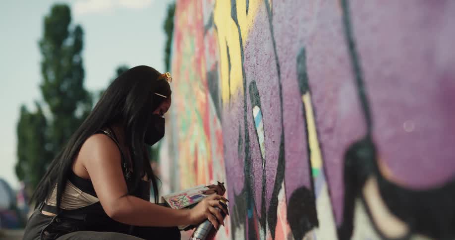 Black woman wearing a face mask using a spray bottle to do graffiti in a big wall outdoors