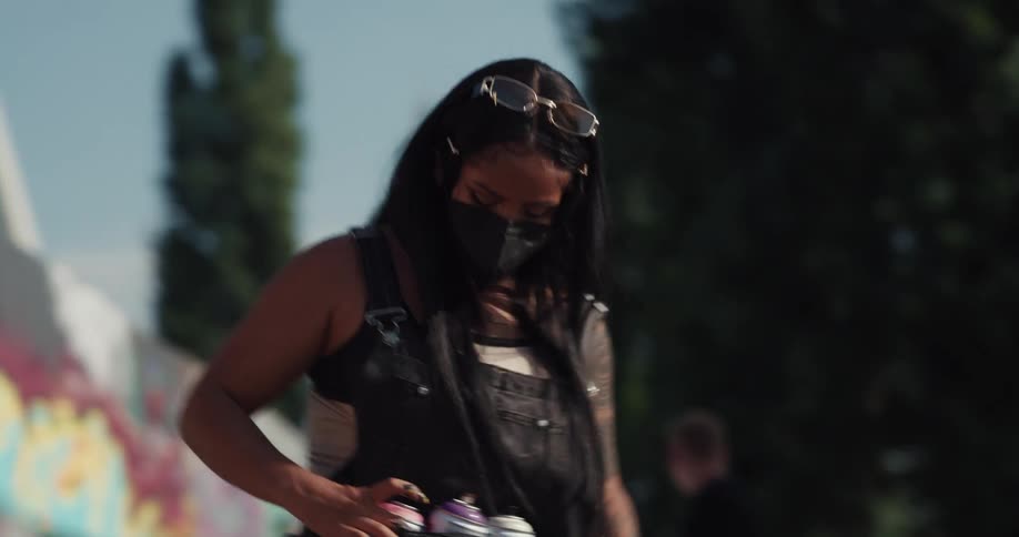 Young woman wearing a black face mask test spray cans before using them to paint graffiti on a wall