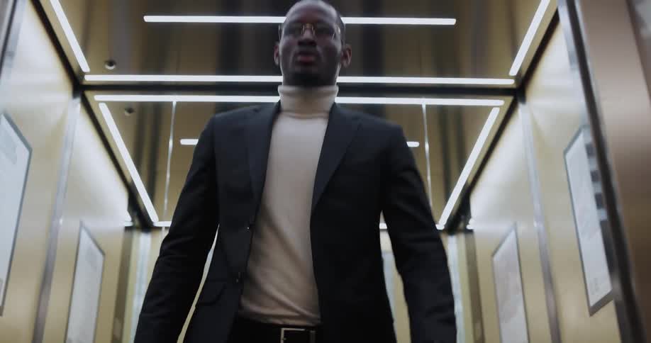 Man wearing a blazer and turtleneck exits an elevator as soon as the doors open