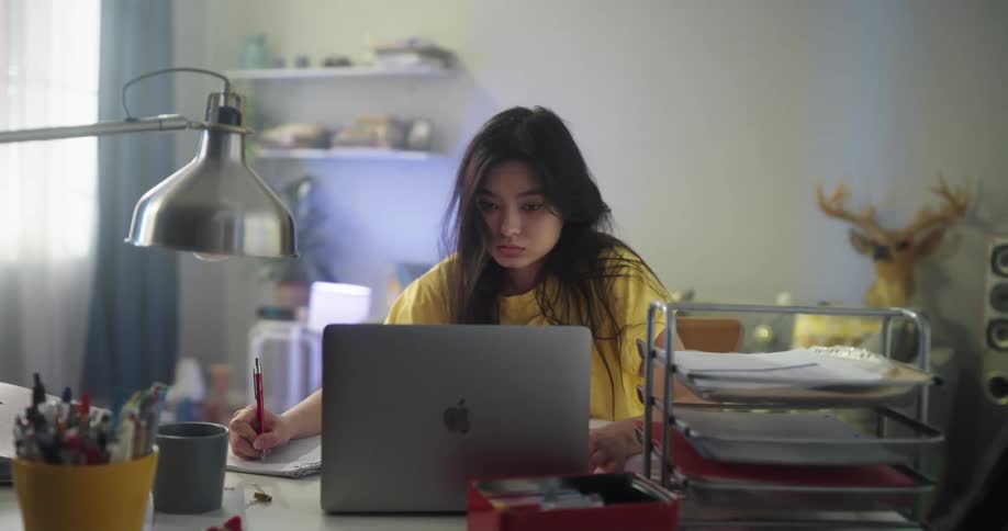 A tired woman opens her eyes wide to try to stay awake as she writes notes while looking at her laptop