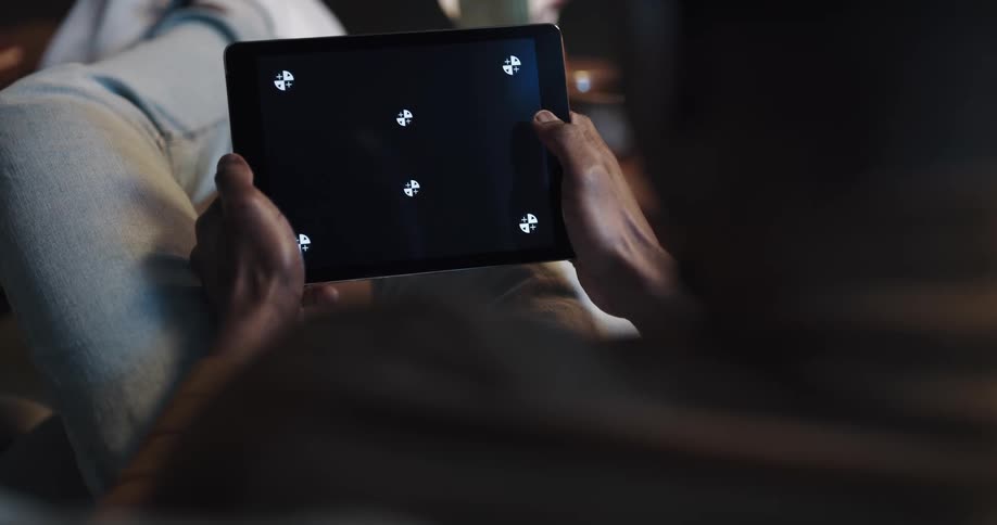 Man with brown turtleneck sweater and jeans sits on couch playing game on tablet