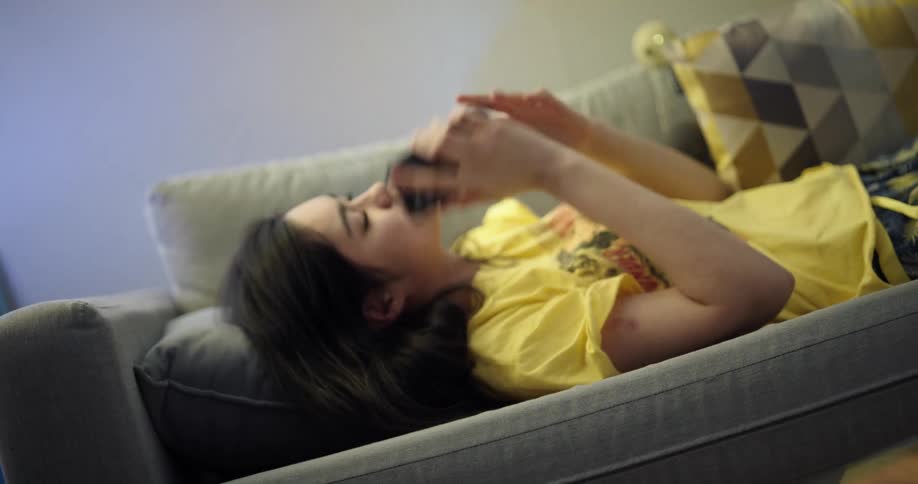 A teenage girl lies on the couch at home and then reaches over to answer her cell phone and talks and laughs