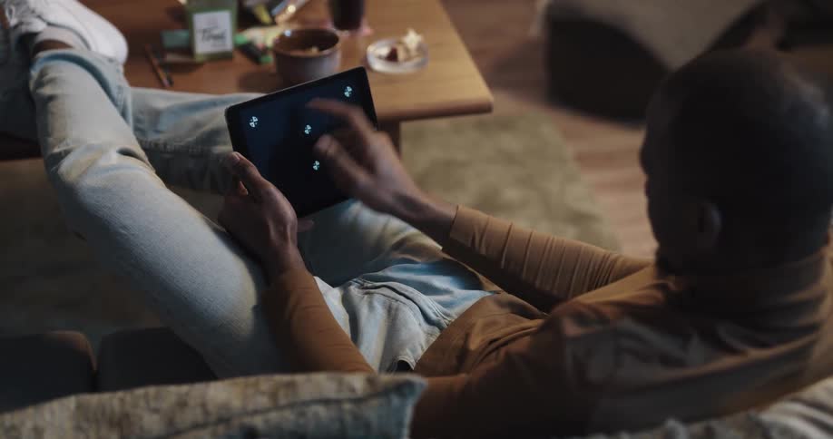 Man with brown turtleneck sweater jeans and white shoes sits on couch and plays with tablet in livng room at home
