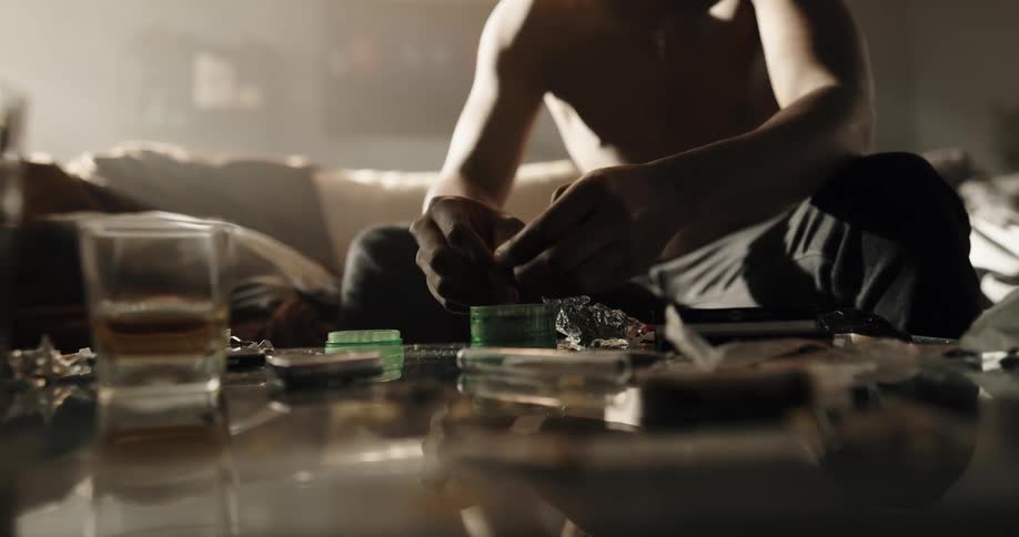 Drug addict sitting on a couch at home fills a handmade cigarette over a coffee table filled with drug paraphernalia