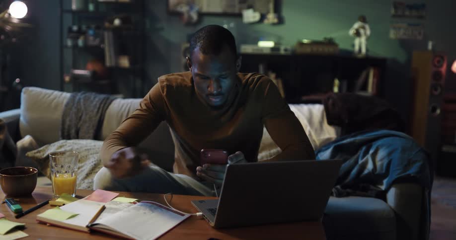 Man with orange sweatshirt and jeans sits on couch using smartphone and laptop in living room at home