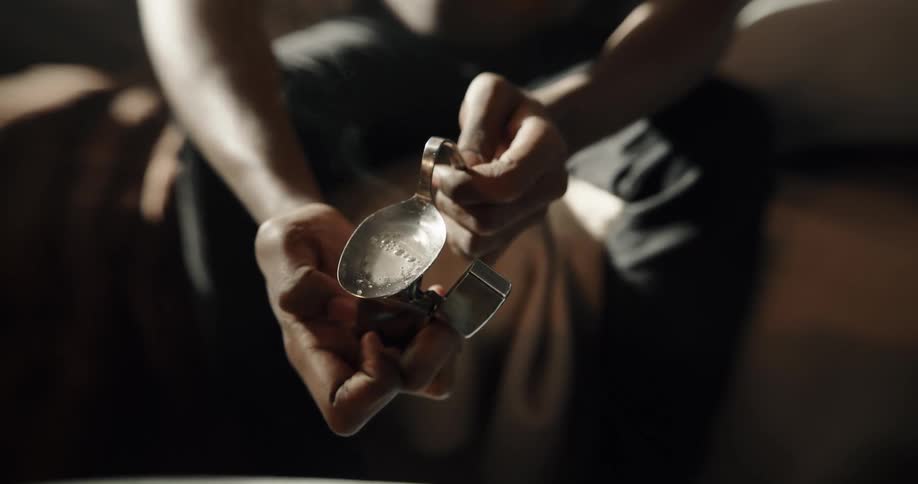 Man places a lighter under a spoon to cook drugs inside a dimly lit room at home