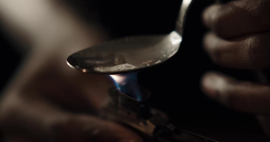 Man places a lighter under a spoon to cook drugs inside a dimly lit room at home