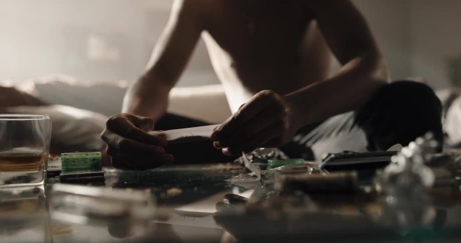 Black man sitting on the couch in front of a table full of drugs and a beverage cup rolling a joint to smoke