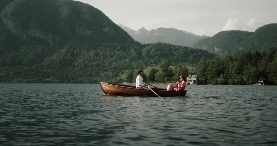 Couple on a date in a wooden canoe in the middle of a lake while the man is rowing and the woman is playing a mini guitar