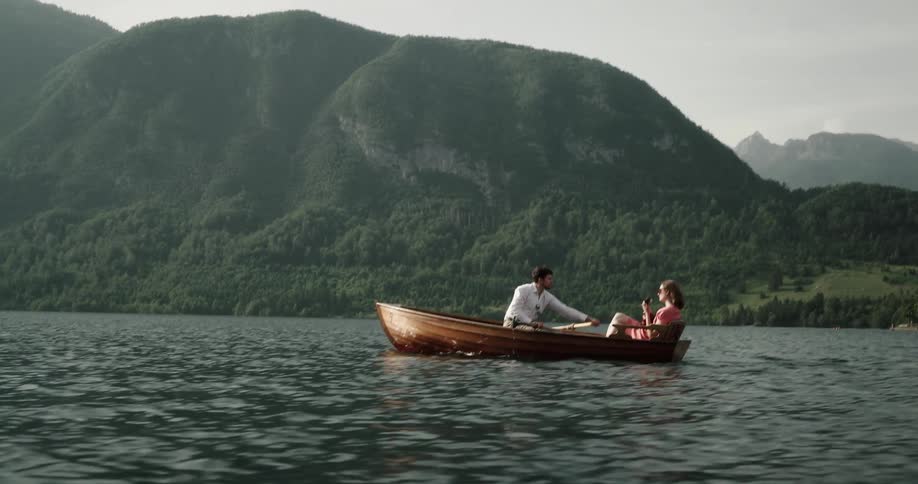 A man and woman are in a small boat on a river near a mountain and he is rowing as she plays a ukulele