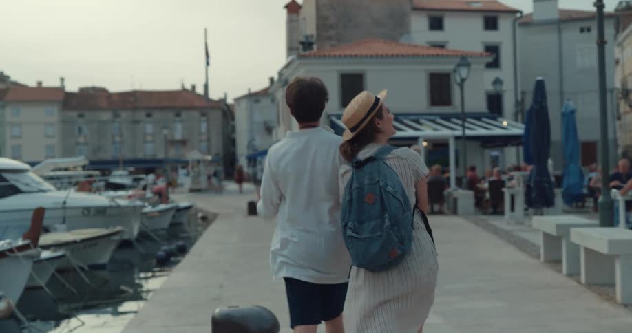 Man with white shirt and black shorts holds womans hand wearing hat backpack and dress walking along promenade and looking at shops in town
