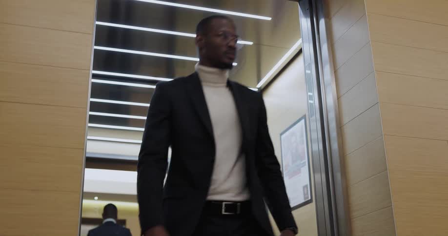 Man wearing a blazer and turtleneck exits an elevator as soon as the doors open