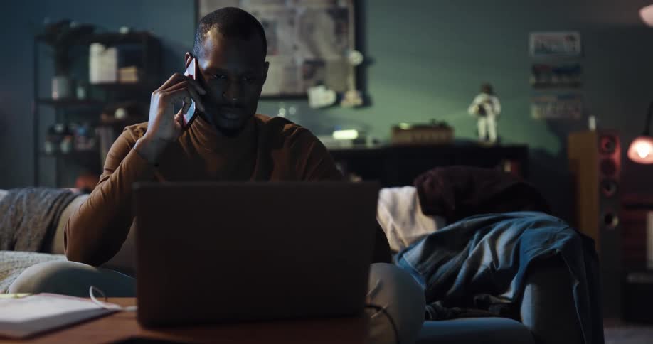Man with brown turtleneck sweater talks on smartphone and uses laptop in living room at home