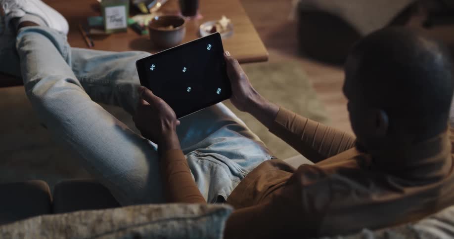 Black man sitting on the couch with his legs up in the center table playing a game on his tablet