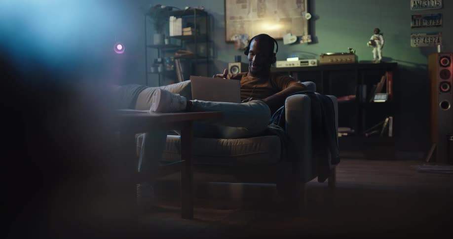 Man with brown turtleneck sweater and jeans sits on couch wearing headphones and using laptop in living room at home