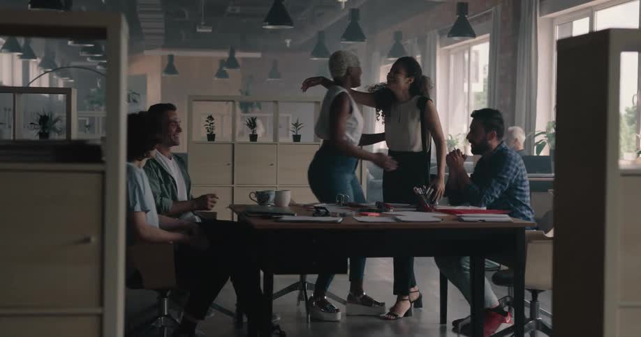 A woman approaches a group of people talking at a table and greets the woman at the table with a hug