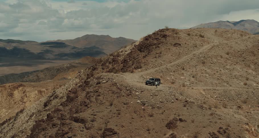 Man walks towards a vehicle that is parked on a dirt road on a mountain under a cloudy sky