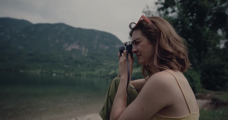 Female photographer sitting on bank near water with mountain in backgroundtakes pictures