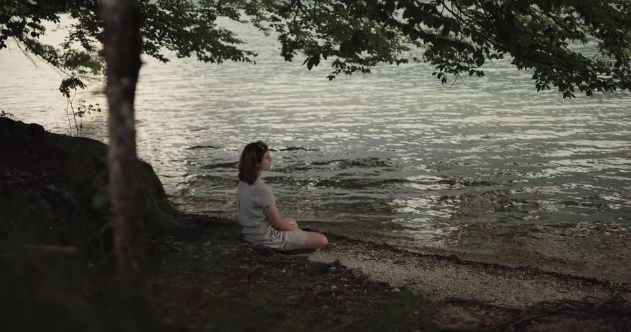 A woman is sitting under a tree by the side of a river as the water ripples in front of her