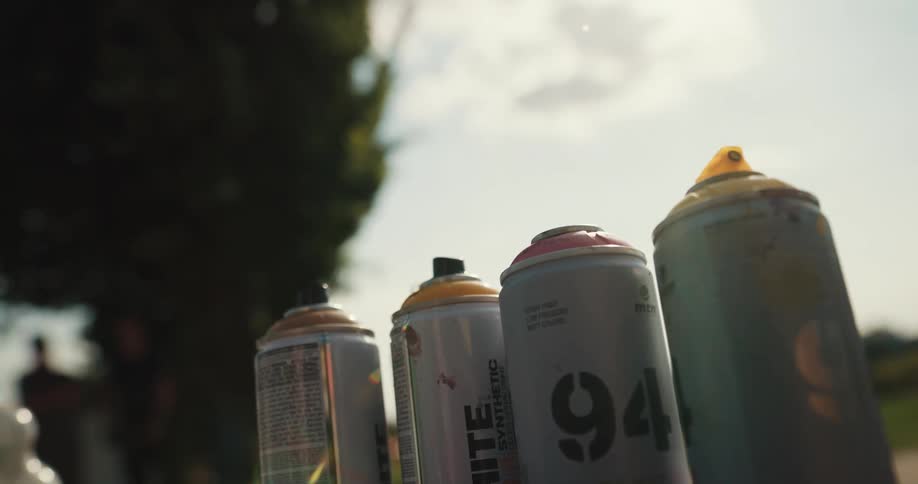 A person picks up a can of spray paint from a group of cans and shakes it while outdoors during the day