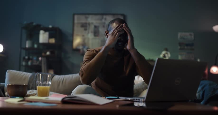 Man with brown turtleck sweater jeans sits on couch using smartphone and laptop and gets frustrated in the living room at home