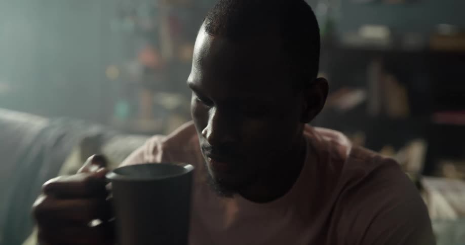 Black man sitting on the couch in the living room drinking a cup of coffee and then standing up