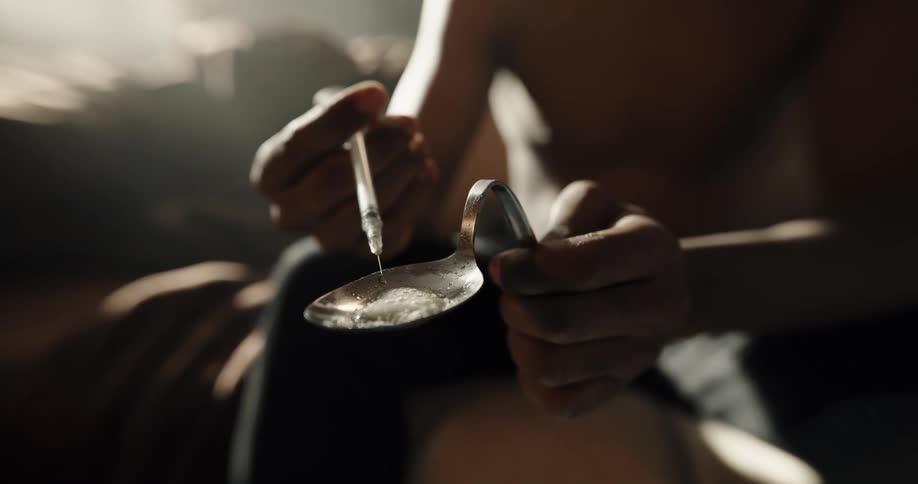 Man mixes liquid from hypodermic with white powder in spoon as he sits in living room