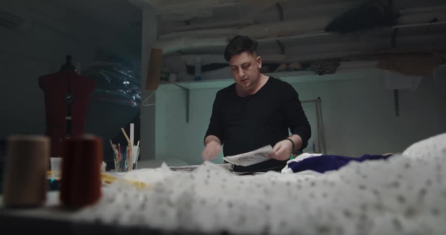 Man walks into dimly lit sewing studio with binder full of designs and starts looking through them