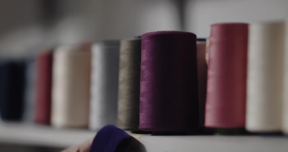 A person compares the color of a fabric to spools of thread on a shelf before retrieving the spool that best matches the color