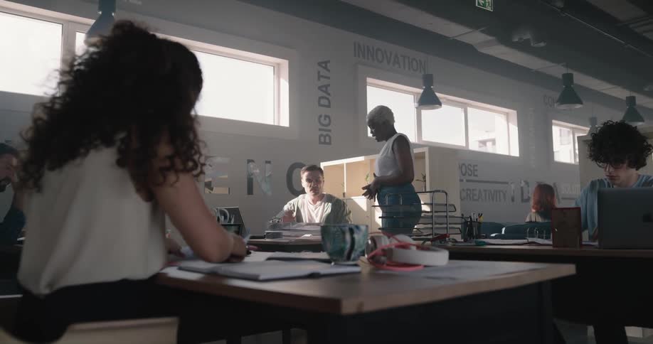 Man sitting at a desk hands a woman a binder while other workers in the office concentrate on their work