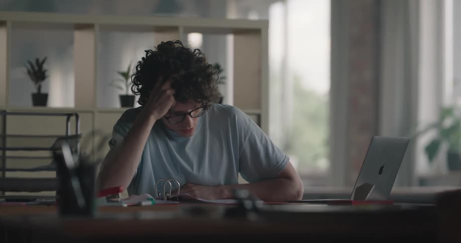 A man is sitting at a desk with a laptop computer and he is looking at pages in a binder