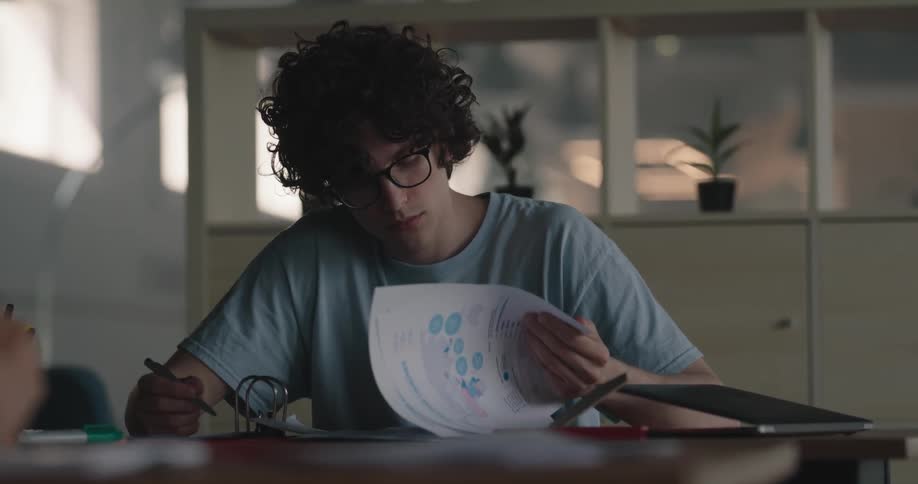 A man is sitting at a desk with a laptop computer and he is looking at pages in a binder