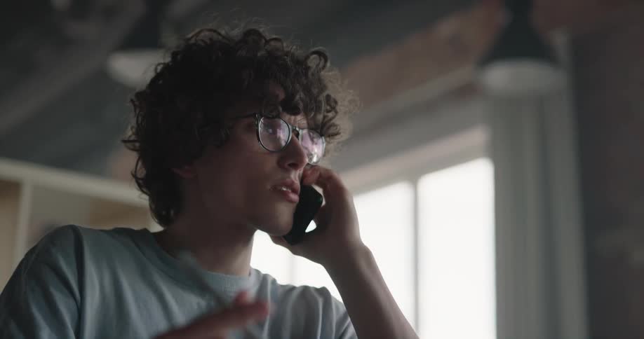 A teenage boy with curly dark hair and glasses is playing with a pen as he talks on the phone