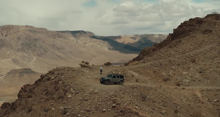 A jeep is parked high on a desert mountain road overlooking a valley and mountains in the distance and a man is walking around