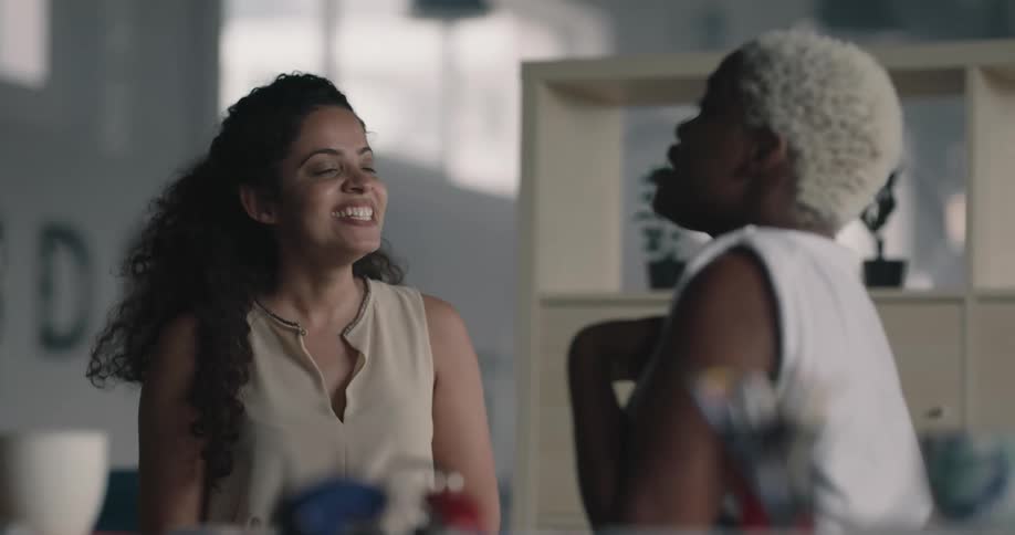 Two female coworkers sitting at the desk in an office space having a conversation and laughing