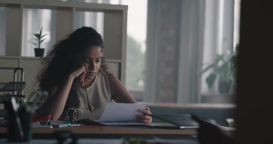 Tired and bored woman sitting on the table at an office space reading some papers