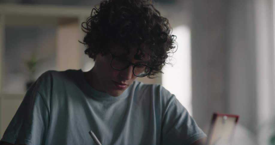 A teenage boy with curly dark hair and glasses is writing on papers