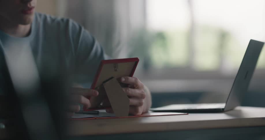 A teenage boy with curly dark hair and glasses is looking at a picture he is holding then he puts it on the desk face down