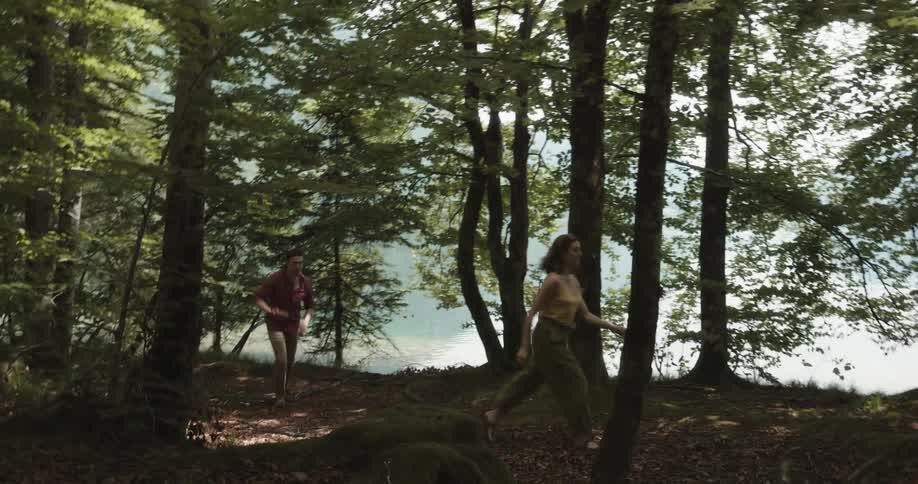 A man chases after a woman as they argue while walking through a woodland next to lake