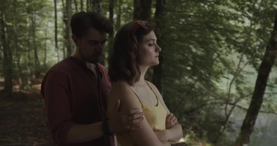 Man with beard and red shirt approaches girl with wavy hair and yellow shirt in the woods next to bench