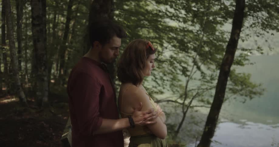 Sad woman standing near a lake with crossed arms while her boyfriend approaches from behind and gently hugs her