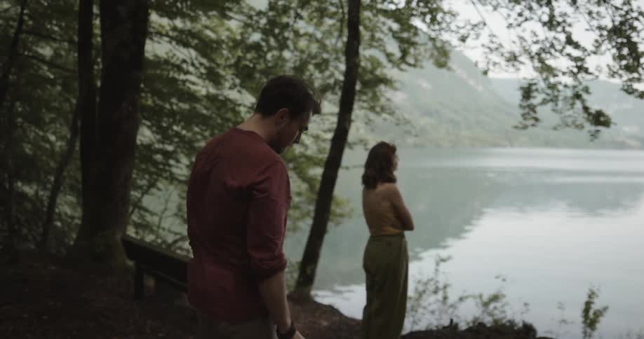 Man removes his hat from his head then nervously approaches a woman standing with her arms crossed in the forest next to the lake