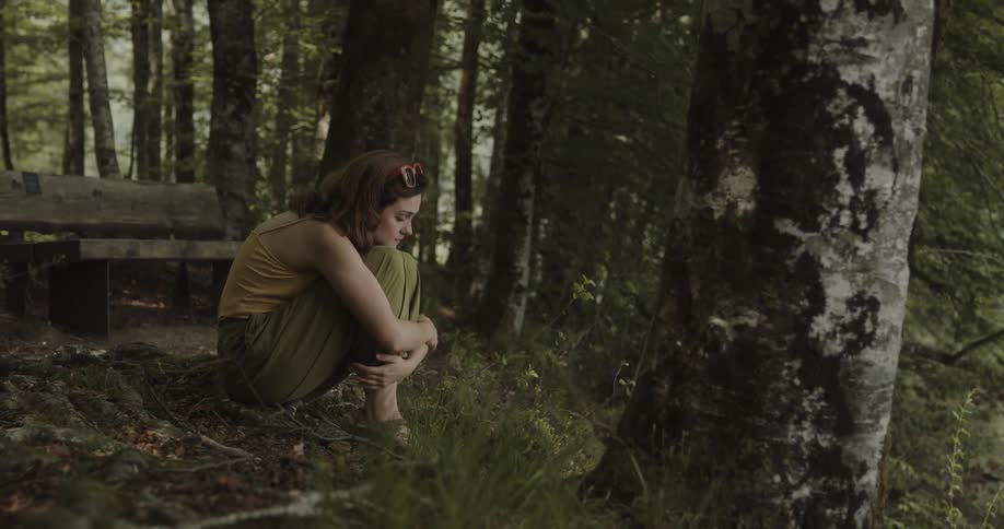 A woman is sitting on a hill in the woods and she appears to be upset