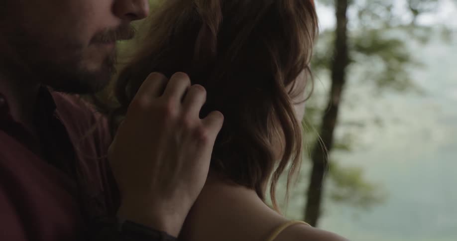 A man stands behind a woman on the shore and plays with her hair and kisses her neck