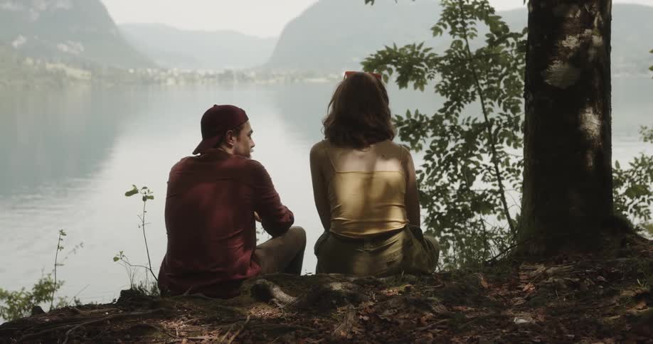 A man approaches a woman looking out over a lake while sitting on the shore and begins to talk to her
