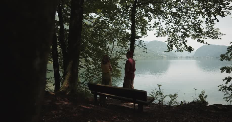 A man walks up to a woman standing in the woods overlooking a lake